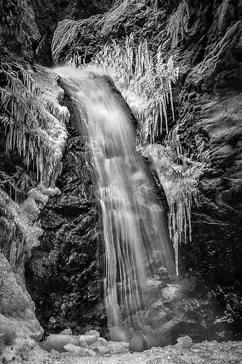 500px provided description: Zapata Falls drops 30 ft / 9 m from an opening at the top of a cave near the base of the Sangre de Cristo Mountains. The opening illuminates the falls and the surrounding ice glows against the dark rock. [#winter ,#water ,#glow ,#rocks ,#black and white ,#monochrome…